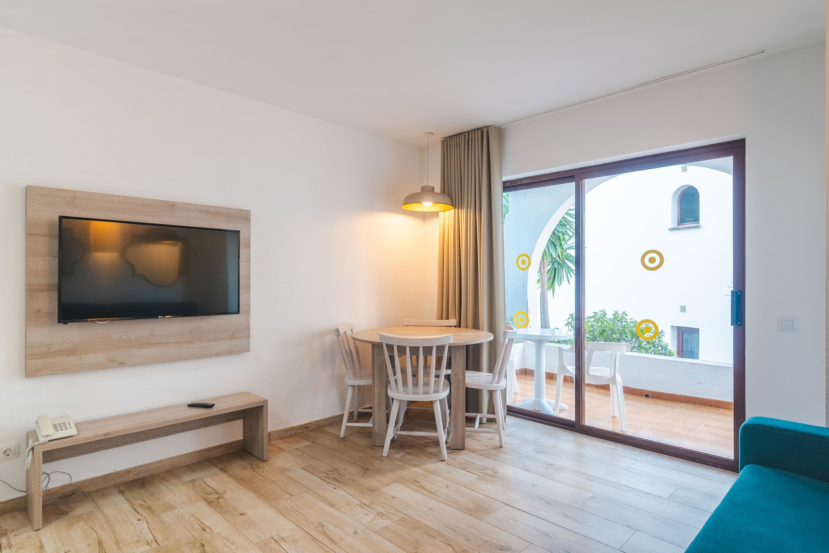 A modern room with a TV mounted on the wall, a round table with chairs, a bench, and a sliding glass door leading to an outdoor area.