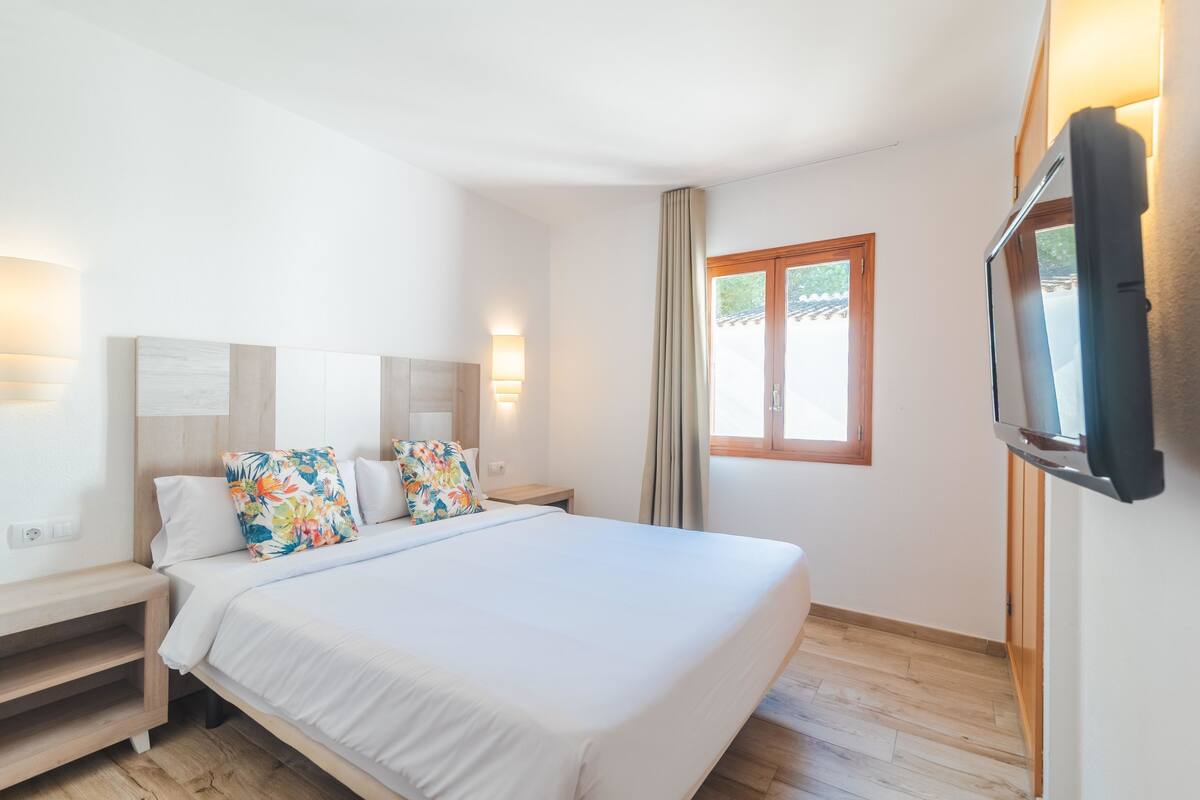 A hotel room with a large bed, a TV, a bedside table, and a window with curtains.