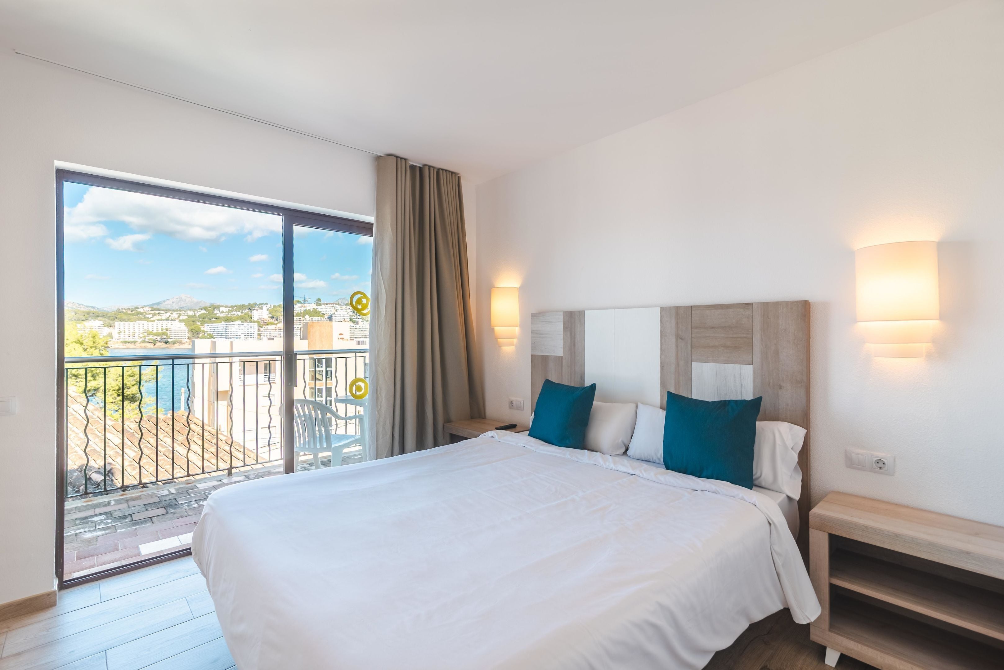 Habitación de hotel con una cama grande, cabecera de madera, dos lámparas de noche y un balcón con vista a la ciudad.