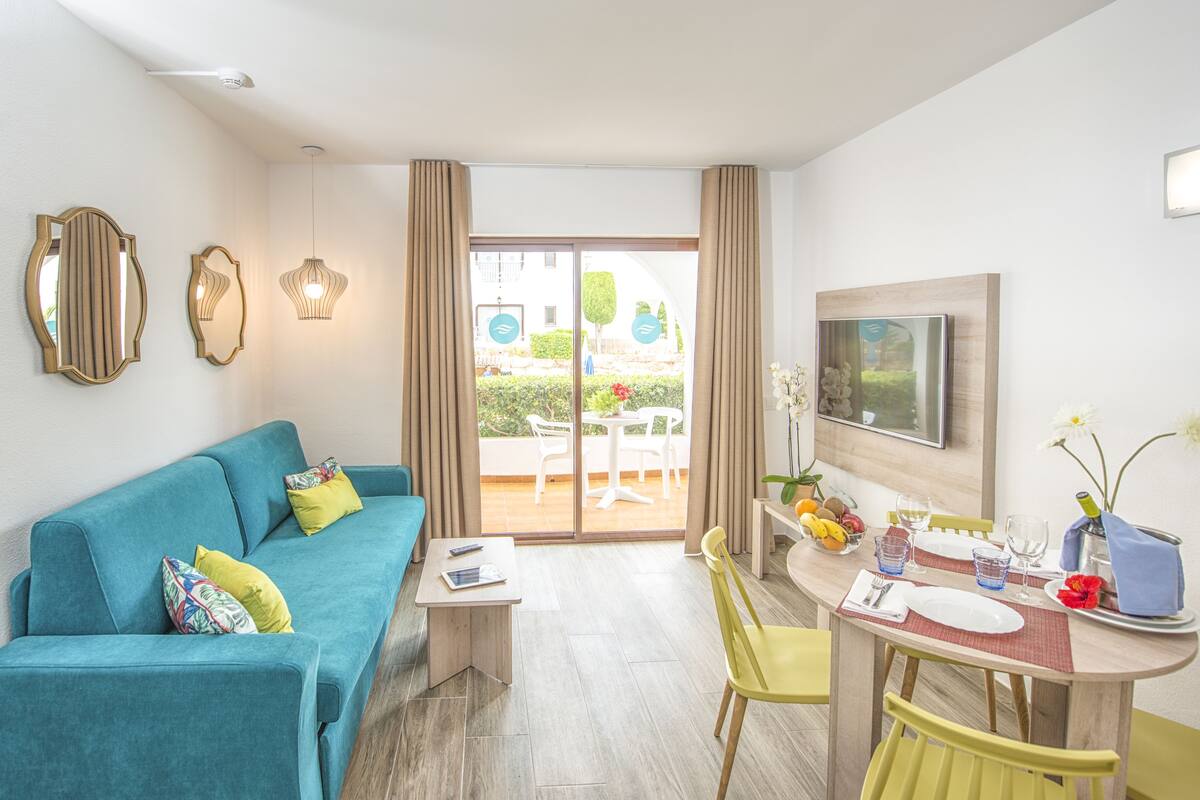 A modern hotel room with a teal sofa, a dining table with yellow chairs, and a balcony with outdoor furniture.