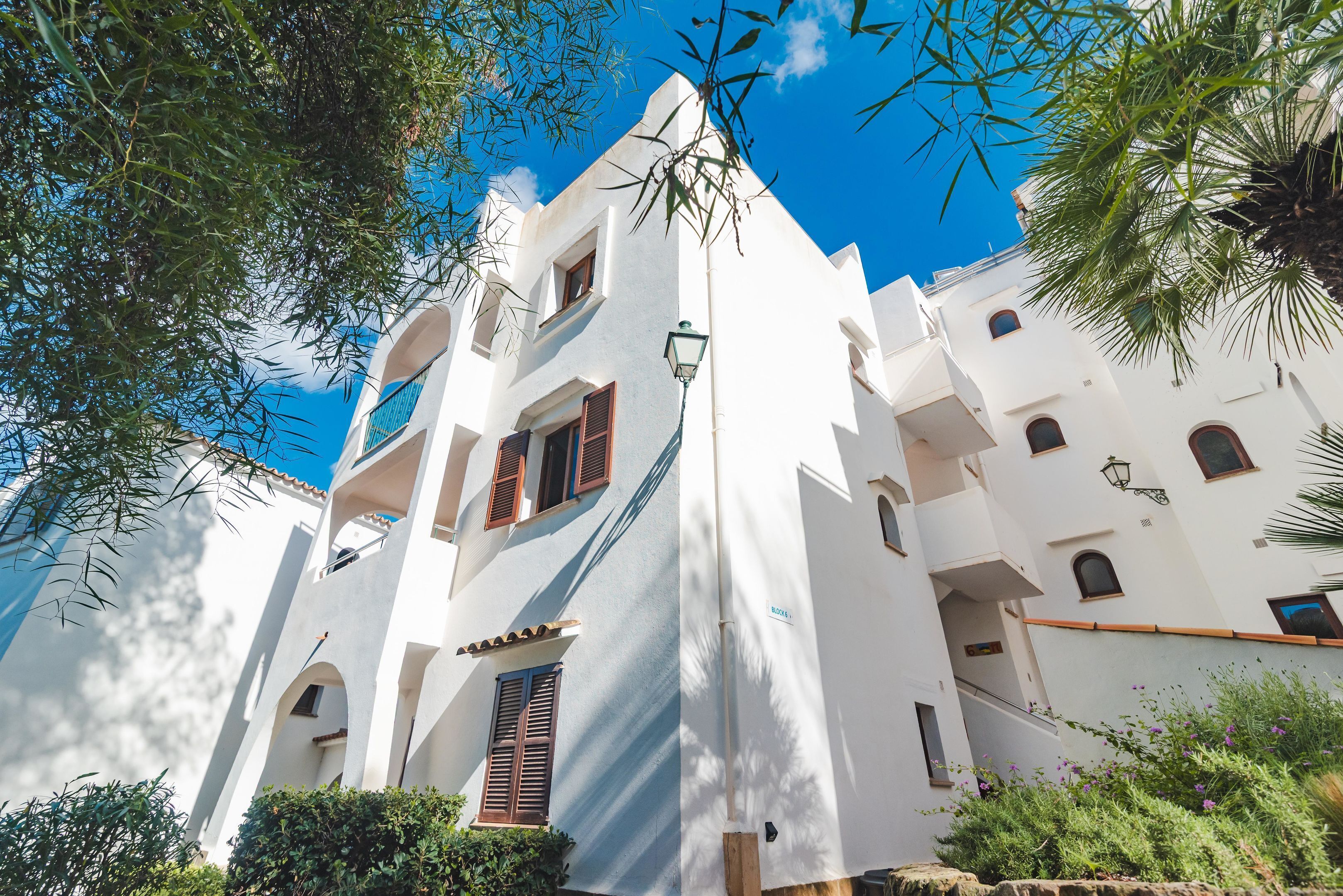 Un edificio blanco con varios balcones y ventanas, rodeado de vegetación y un cielo azul despejado.