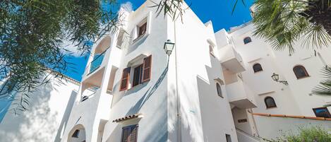 Un edificio blanco con varios balcones y ventanas, rodeado de vegetación y un cielo azul despejado.