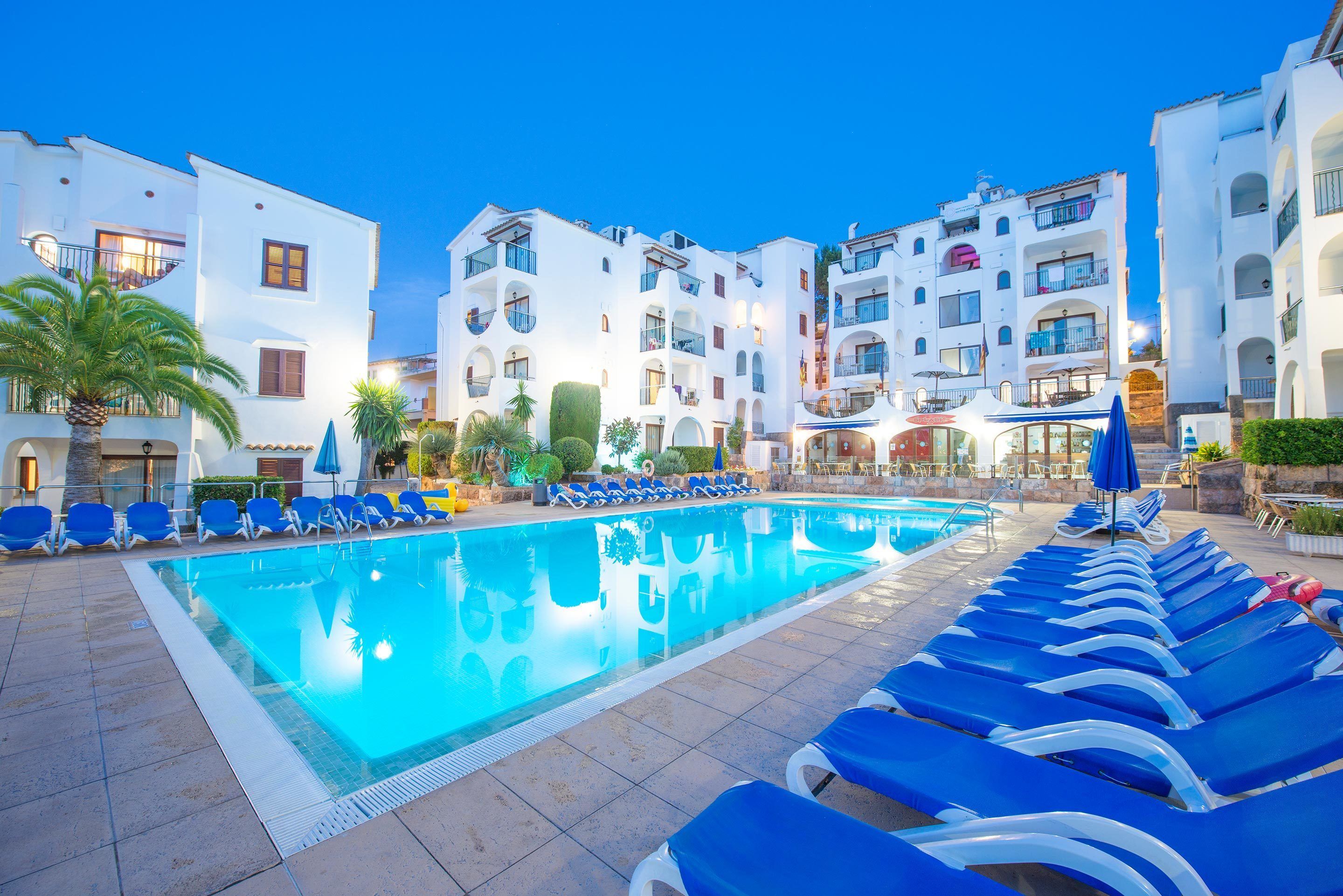 A swimming pool with blue lounge chairs, white buildings, and a clear sky.