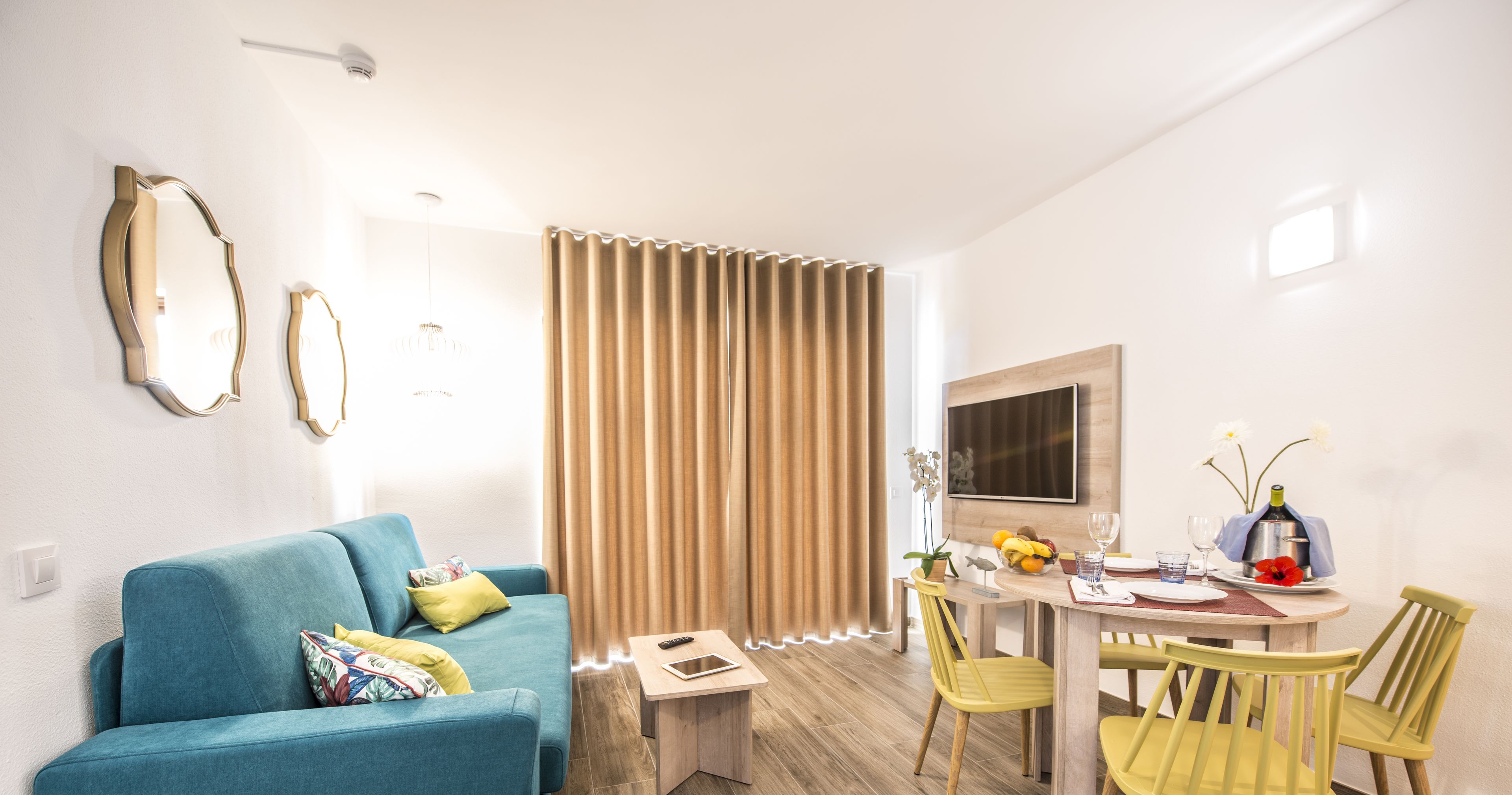 Un salon moderne avec un canapé bleu, une table à manger avec des chaises jaunes, un téléviseur fixé au mur et une fenêtre avec des rideaux.