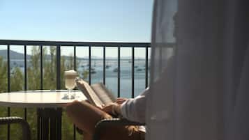A person sitting at a table with a book and a glass of water, overlooking a body of water with boats and distant land.