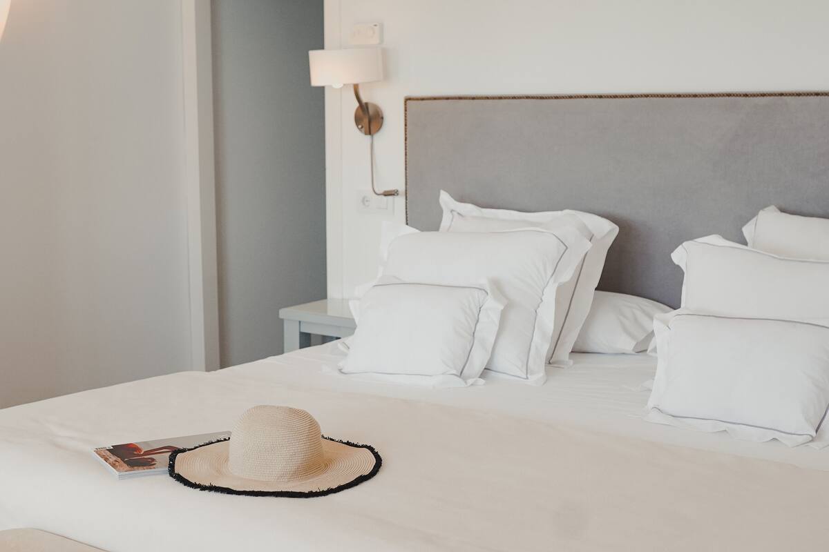 Un lit bien fait, avec du linge de lit blanc et des oreillers, une petite table de chevet avec une lampe, et un chapeau de paille posé sur le lit.