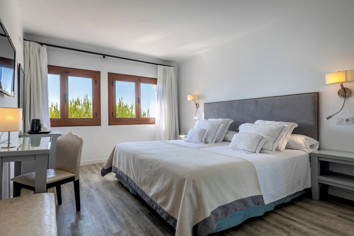Un dormitorio con una cama grande, un escritorio y una silla. Hay dos ventanas con cortinas, una lámpara en la pared y vistas al exterior con vegetación.
