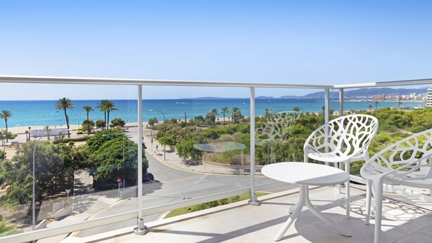 Un balcón con sillas blancas y una mesa, con vista a una playa con palmeras y un cielo azul despejado.