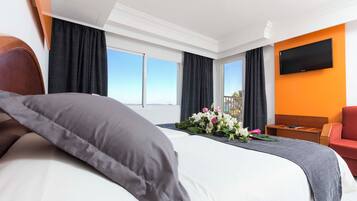 A hotel room with a large bed, a flat-screen TV mounted on the wall, and a view of the sky through the windows.