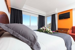 A hotel room with a large bed, a flat-screen TV mounted on the wall, and a view of the sky through the windows.