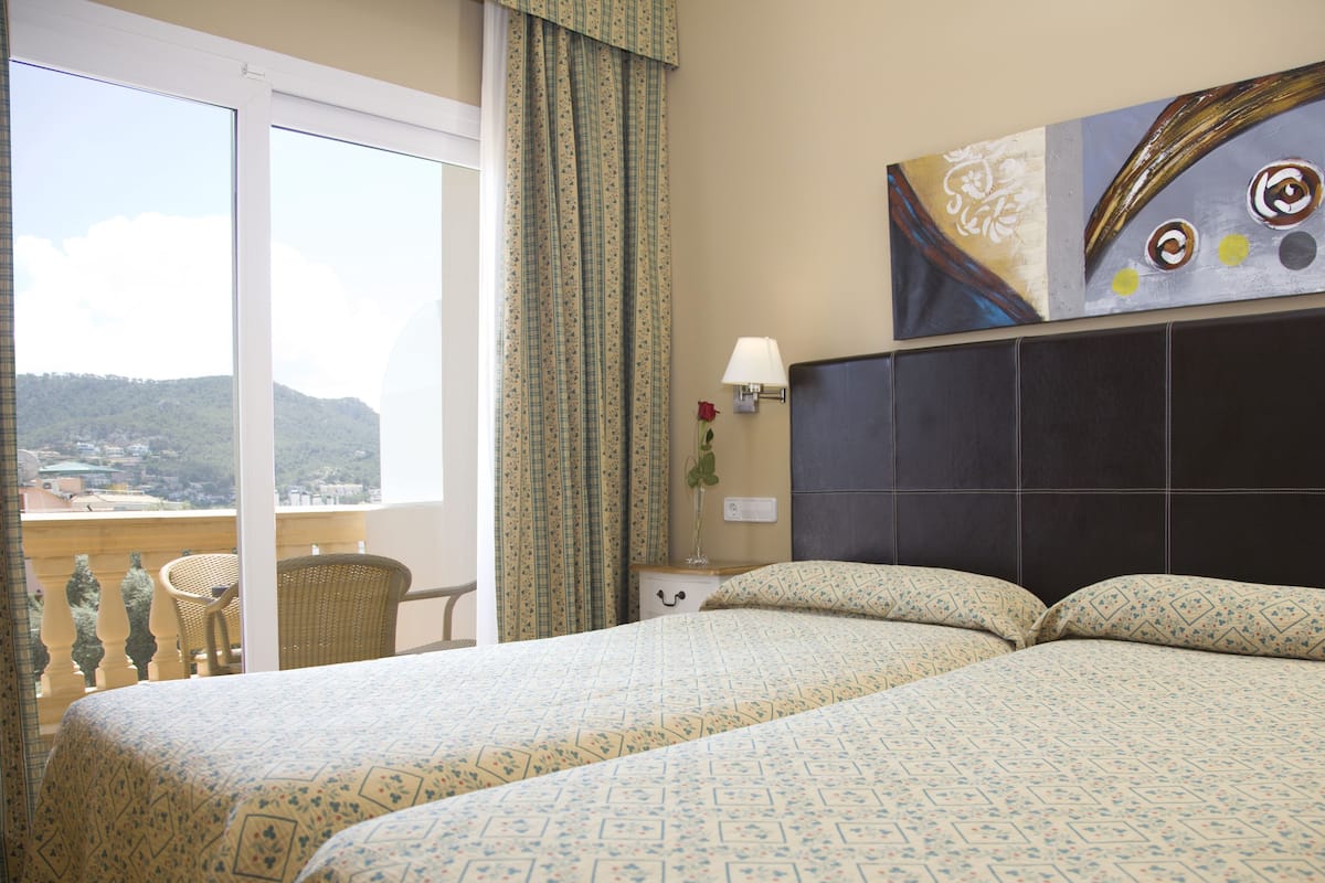 Habitación de hotel con dos camas, un ventanal con vistas a la montaña, un cuadro en la pared y un jarrón con una rosa.