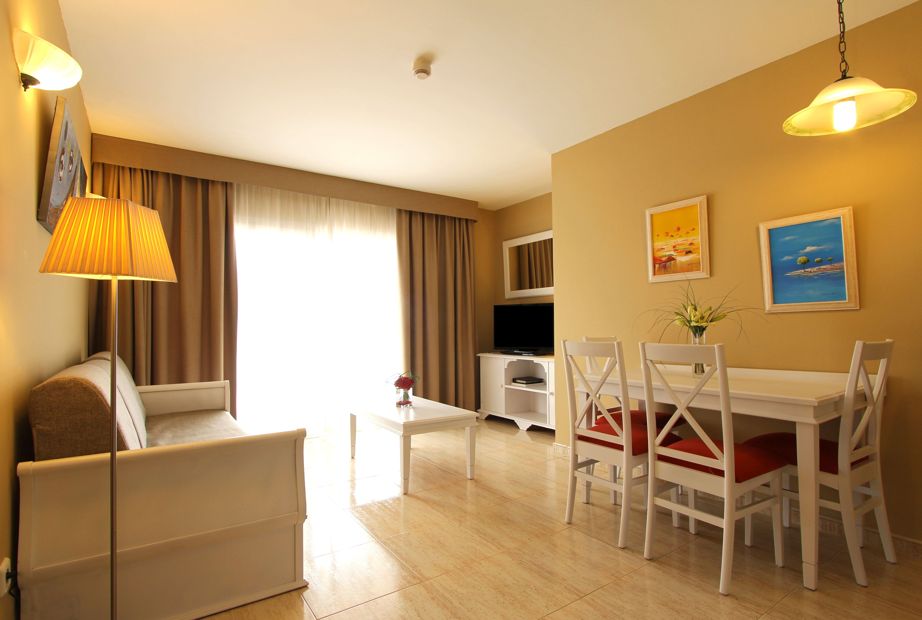 A hotel room with a sofa, dining table, chairs, a TV, and artwork on the walls.