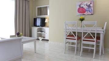 Une chambre d’hôtel dotée d’un coin repas comprenant une table et des chaises blanches, un téléviseur posé sur un meuble et un tableau accroché au mur.