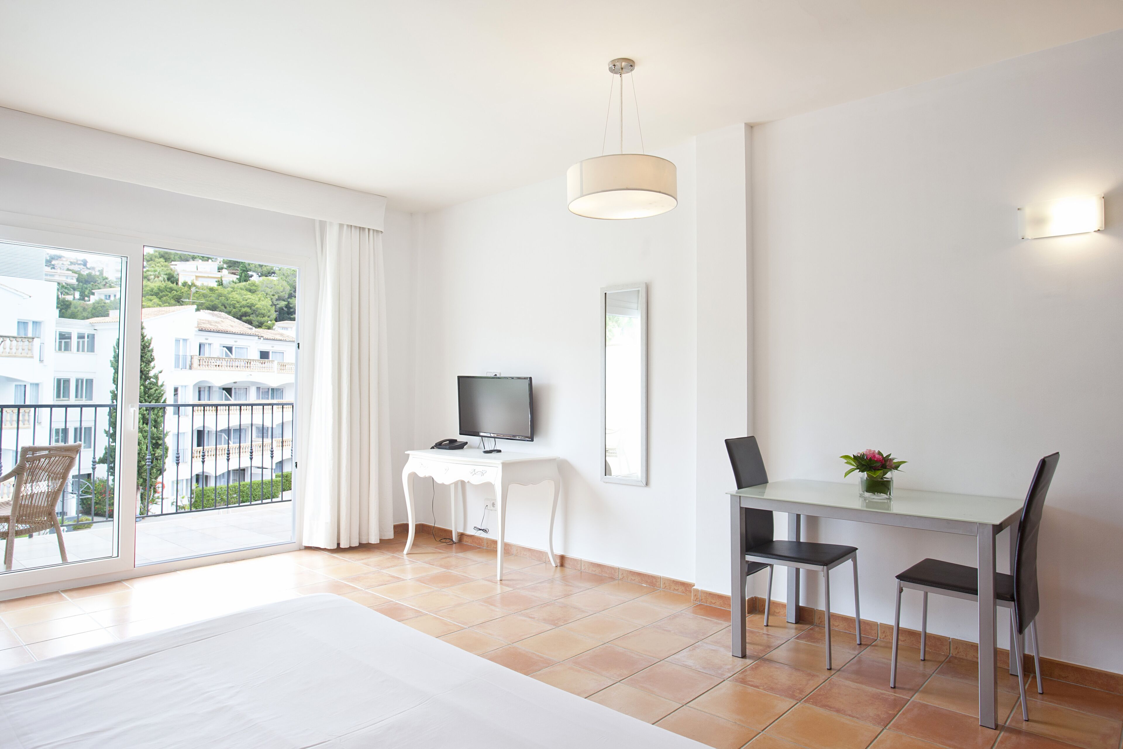 A modern hotel room with a balcony, a dining area with a table and chairs, a TV on a white console table, and a large window with curtains.