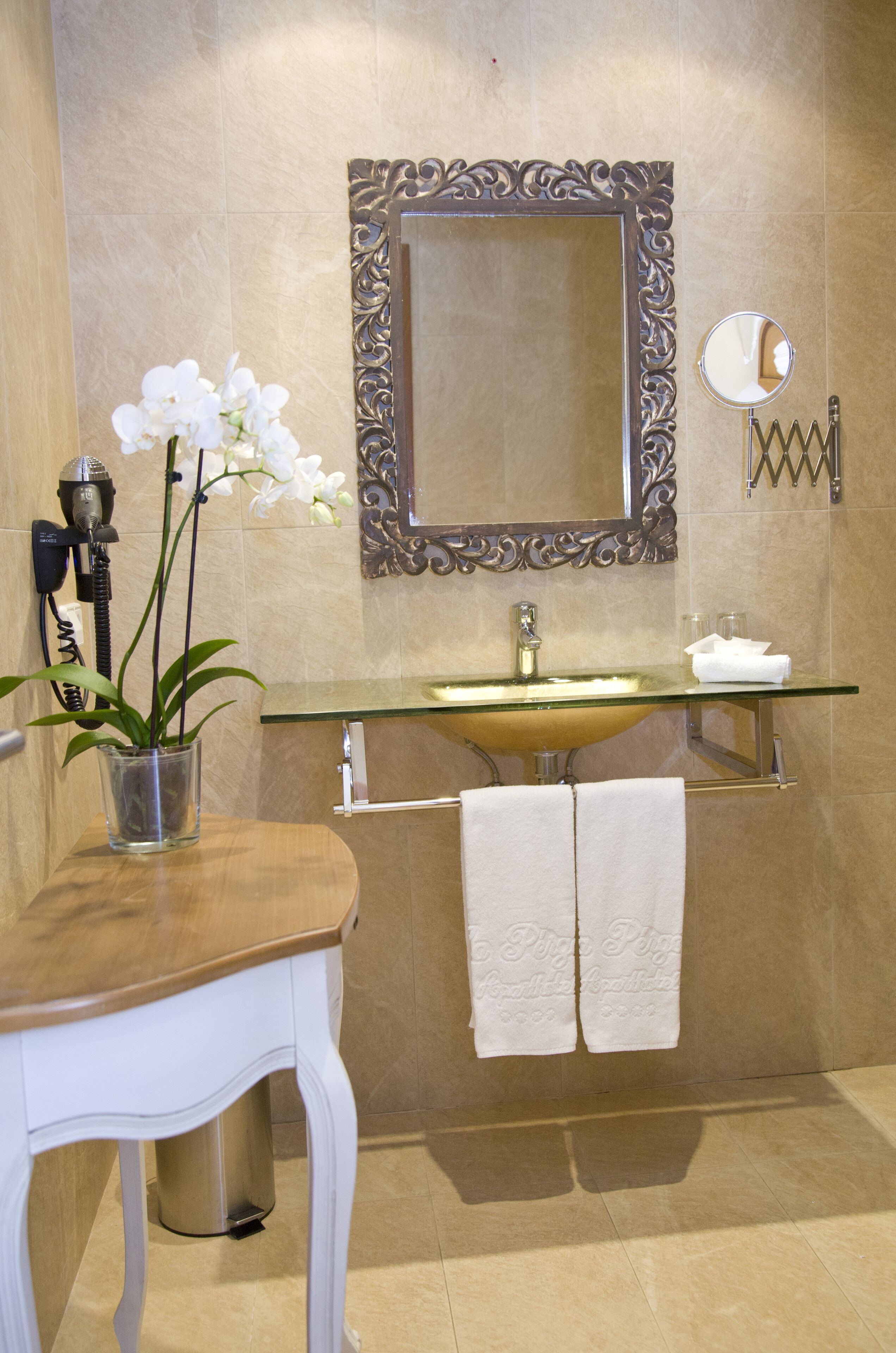 Een badkamer met een glazen wastafel, een spiegel met een sierlijk frame, een potorkidee en twee handdoeken aan de muur.