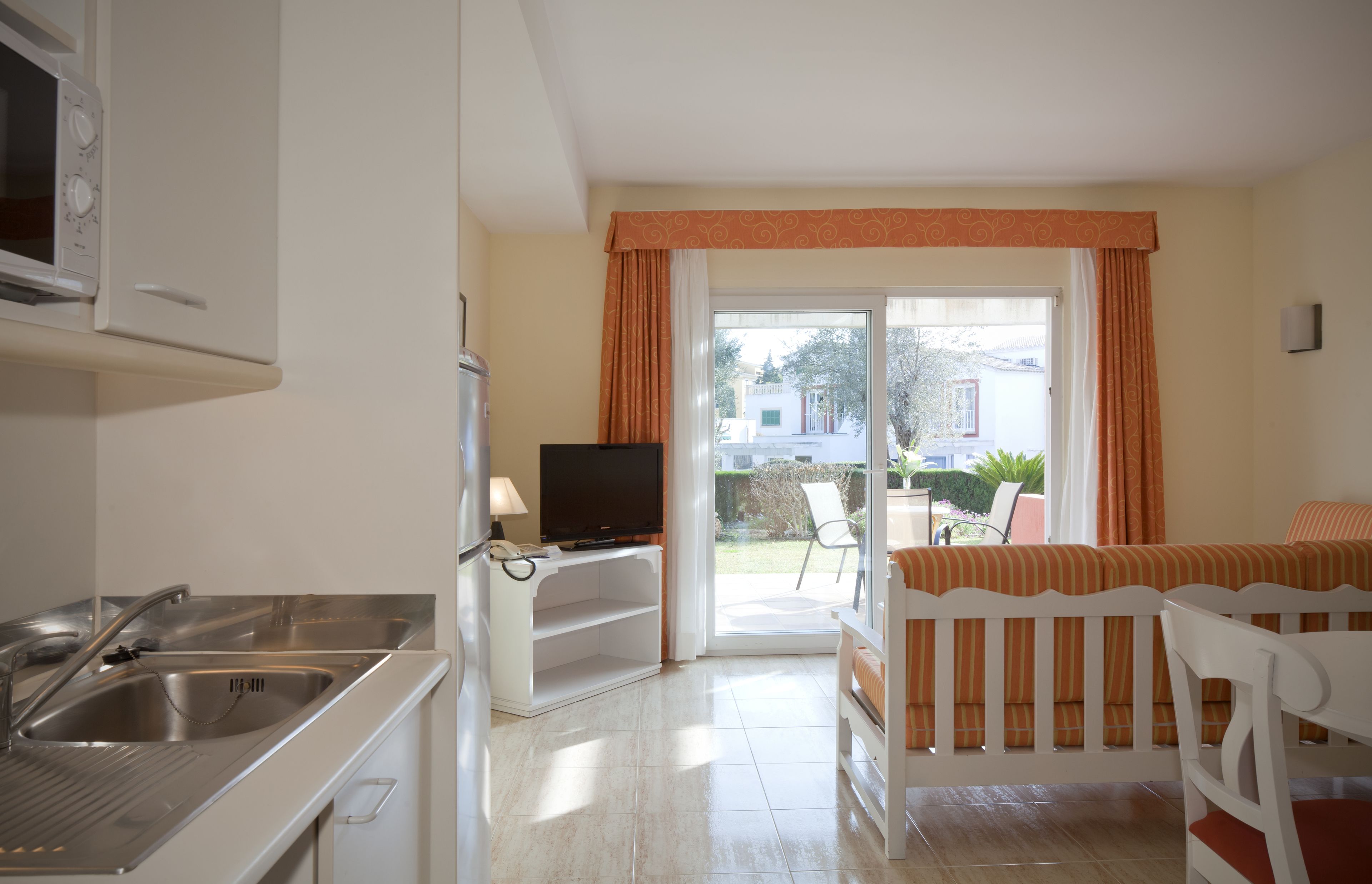 Una cocina con microondas, lavamanos y televisor. Hay un balcón con una silla y una mesa.