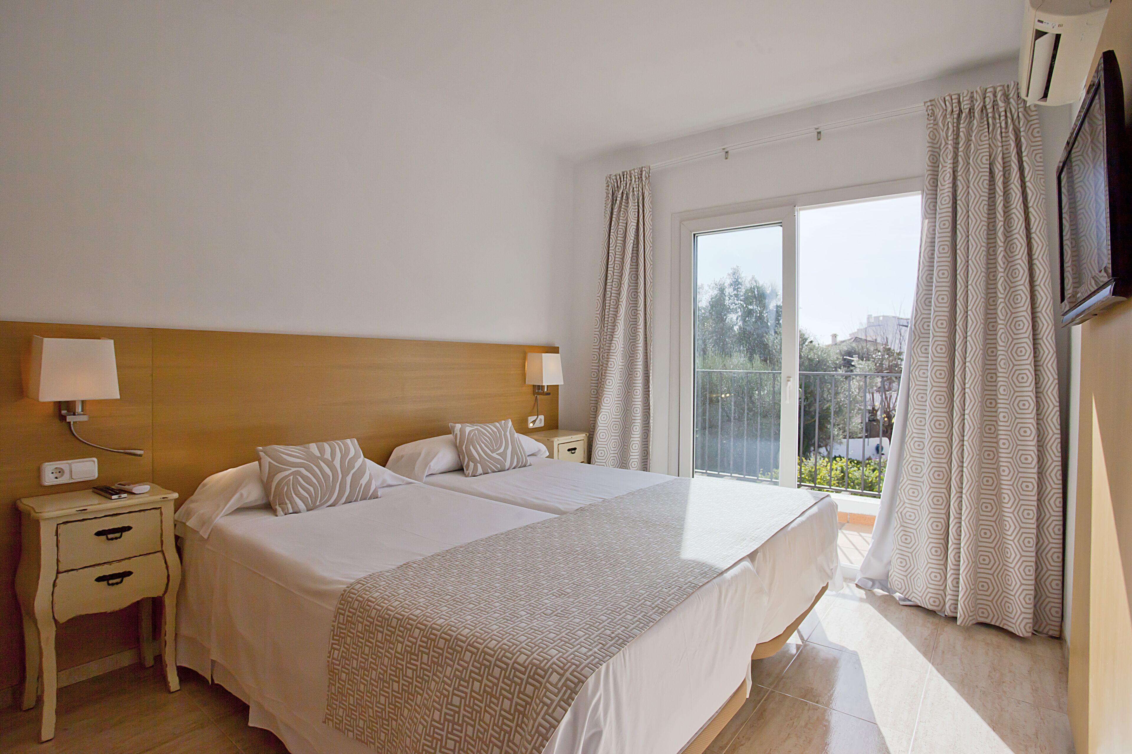 A hotel room with two beds, a wooden headboard, a nightstand with a lamp, and a large window with curtains.