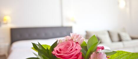 A bed with white bedding and pillows, a bedside table with a lamp, and a bouquet of pink flowers.