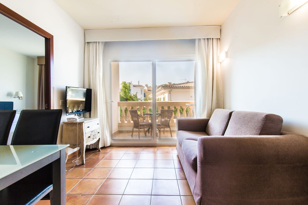 Un salón con sofá, televisor sobre un mueble bajo, mesa de comedor y balcón con vistas a edificios.