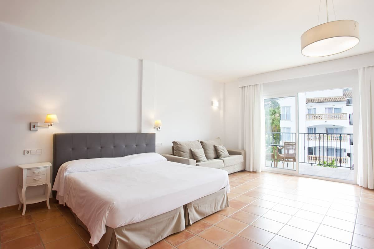 Un dormitorio con una cama grande, una mesita de noche, un sofá y un balcón con vistas a edificios.