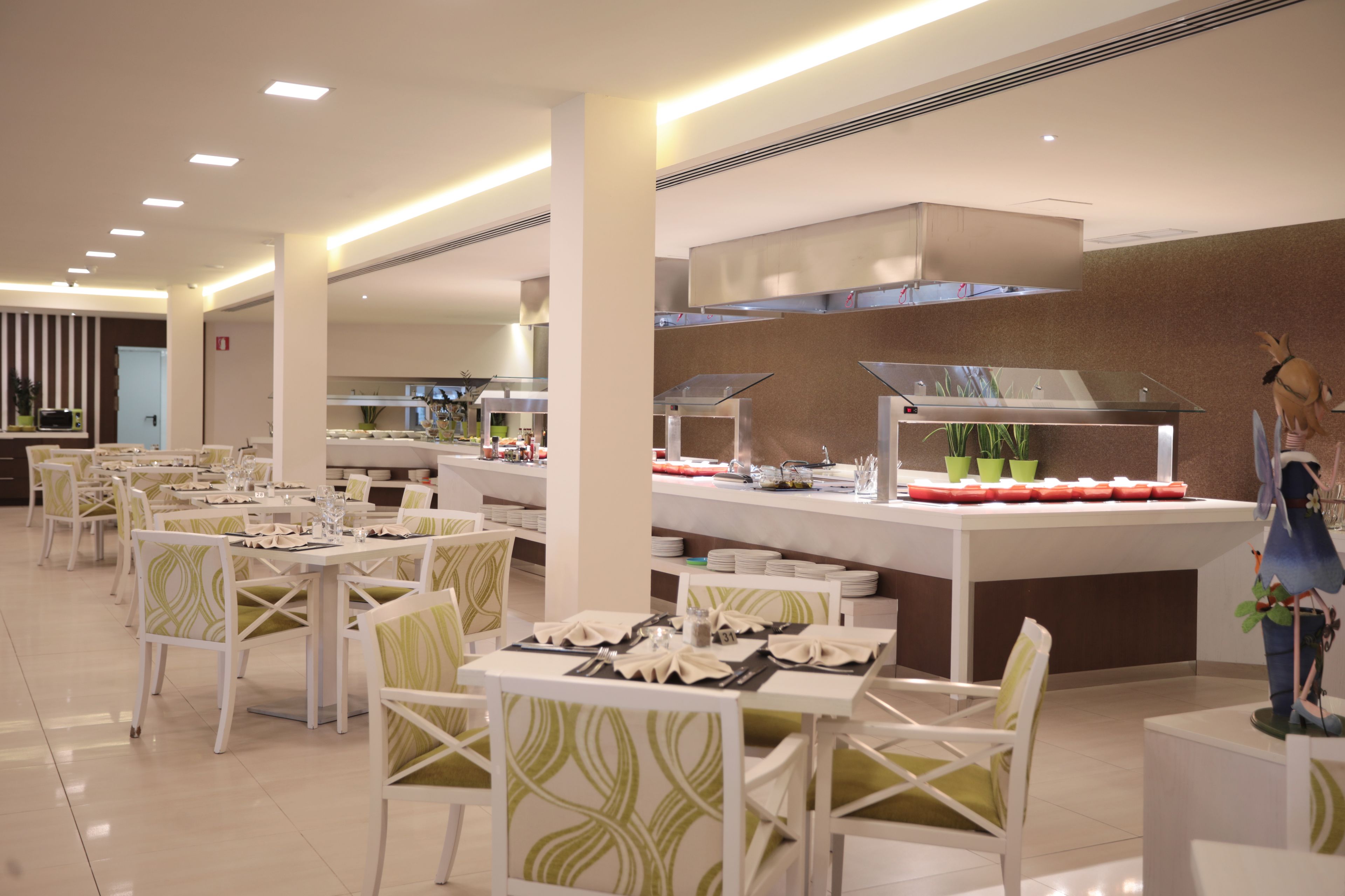 A modern dining area with white tables and chairs, a buffet counter with food displays, and a decorative statue.