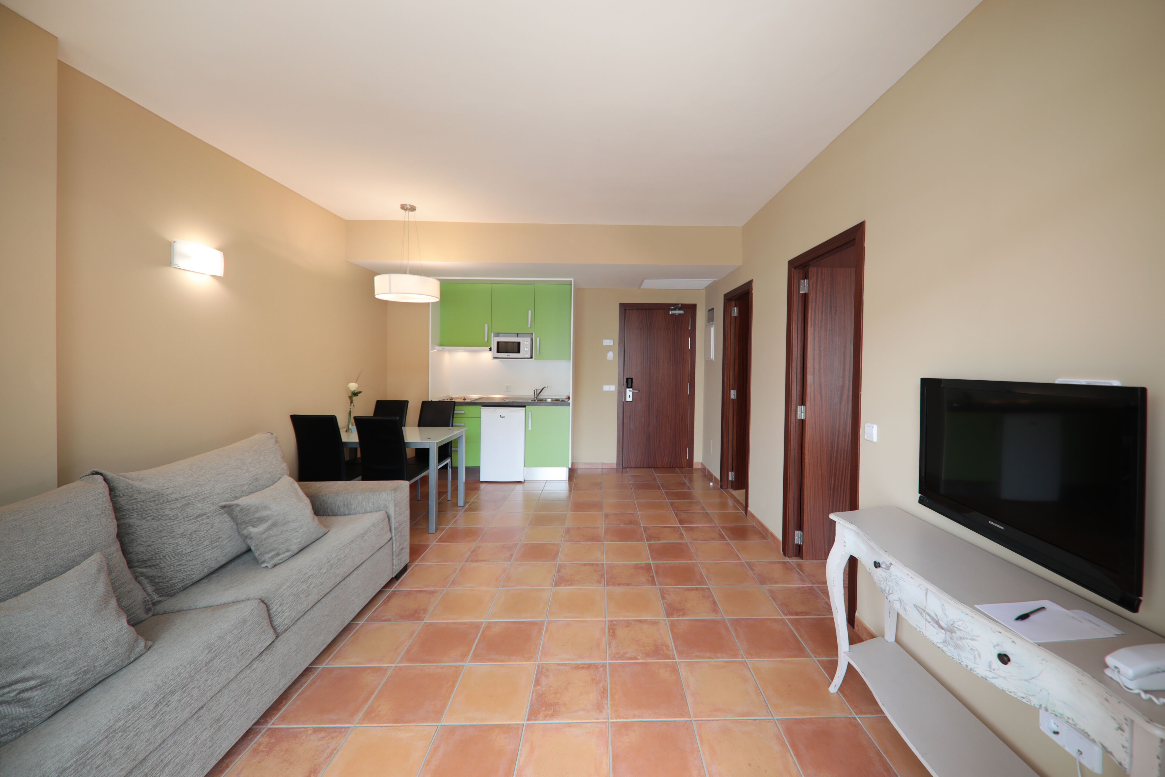 A living room with a grey sofa, a television on a white stand, and a kitchen area in the background.