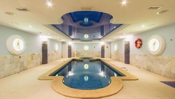 A large indoor swimming pool with a central circular feature, surrounded by a tiled floor and walls with circular windows.