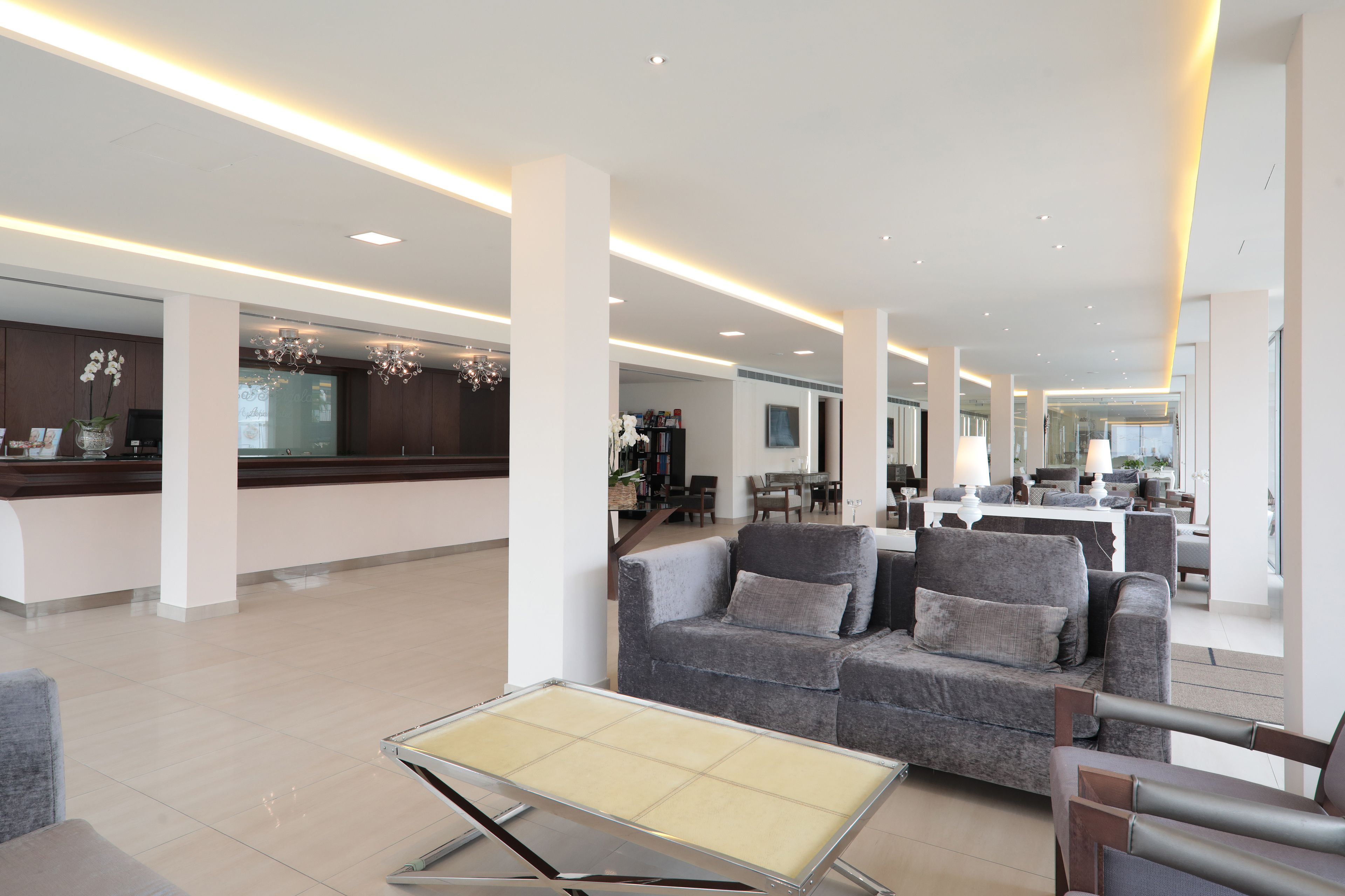 A spacious lobby with a grey sofa, a small table, and a reception area with a counter and decorative plants.