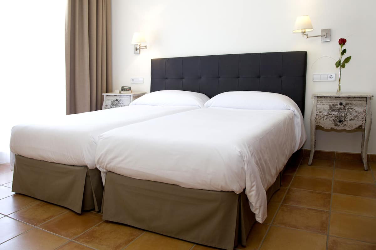 Una cama doble con cabecera oscura, dos mesitas de noche con lámparas, un florero con una rosa y una cortina en la ventana.