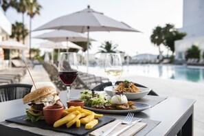 Una mesa de comedor con una hamburguesa, papas fritas, ensalada y dos copas de vino, con una piscina y sombrillas al fondo.