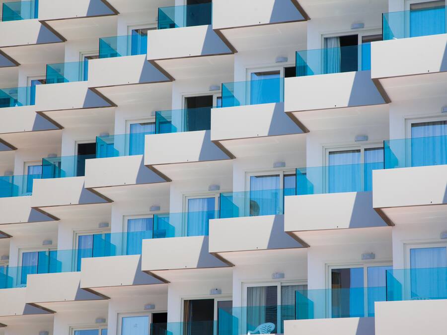 Un edificio moderno con fachada geométrica, que presenta balcones con barandillas de vidrio y numerosas ventanas.