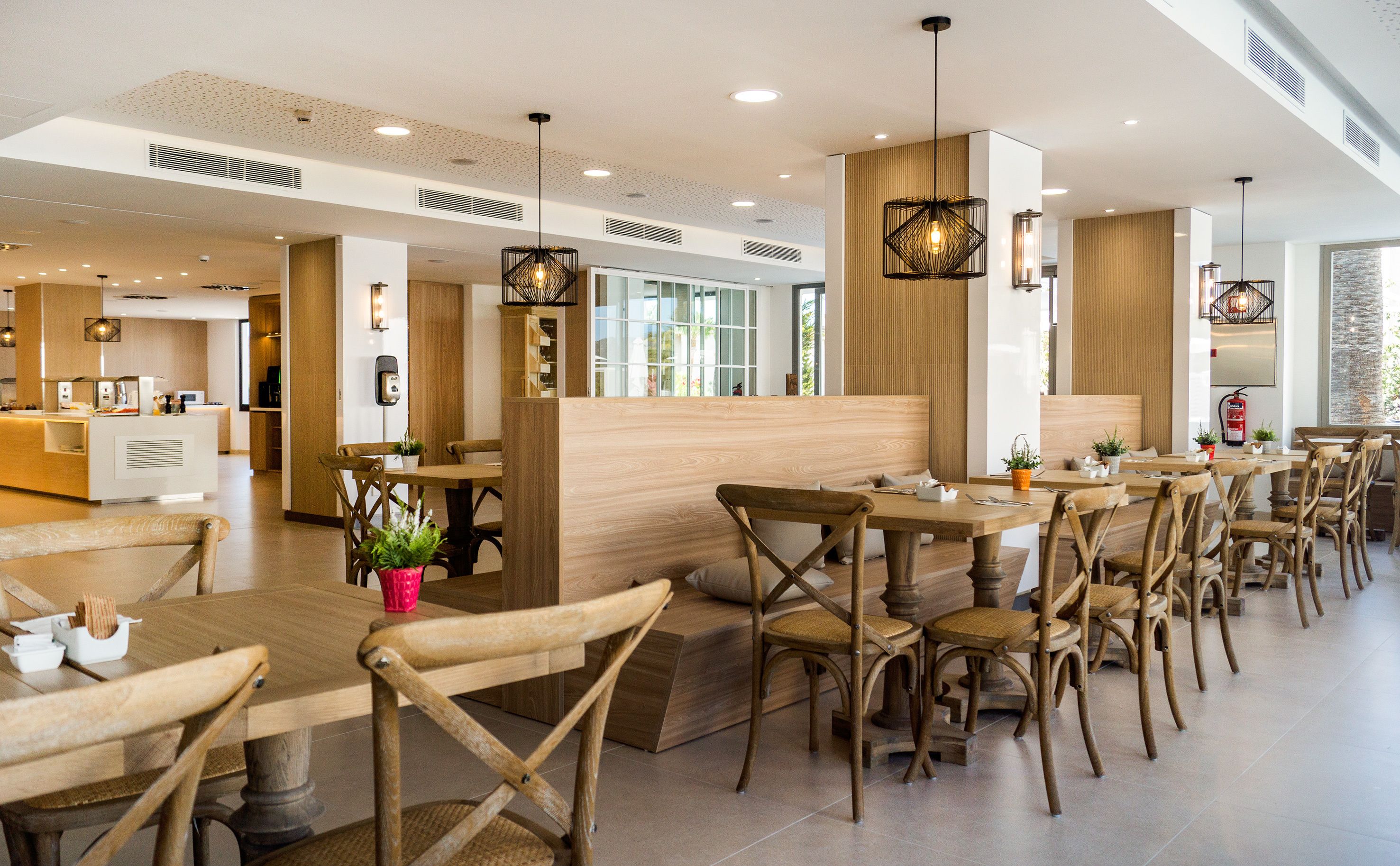 L’intérieur d’un restaurant moderne, avec des tables et des chaises en bois, des luminaires suspendus et un espace cuisine en arrière-plan.