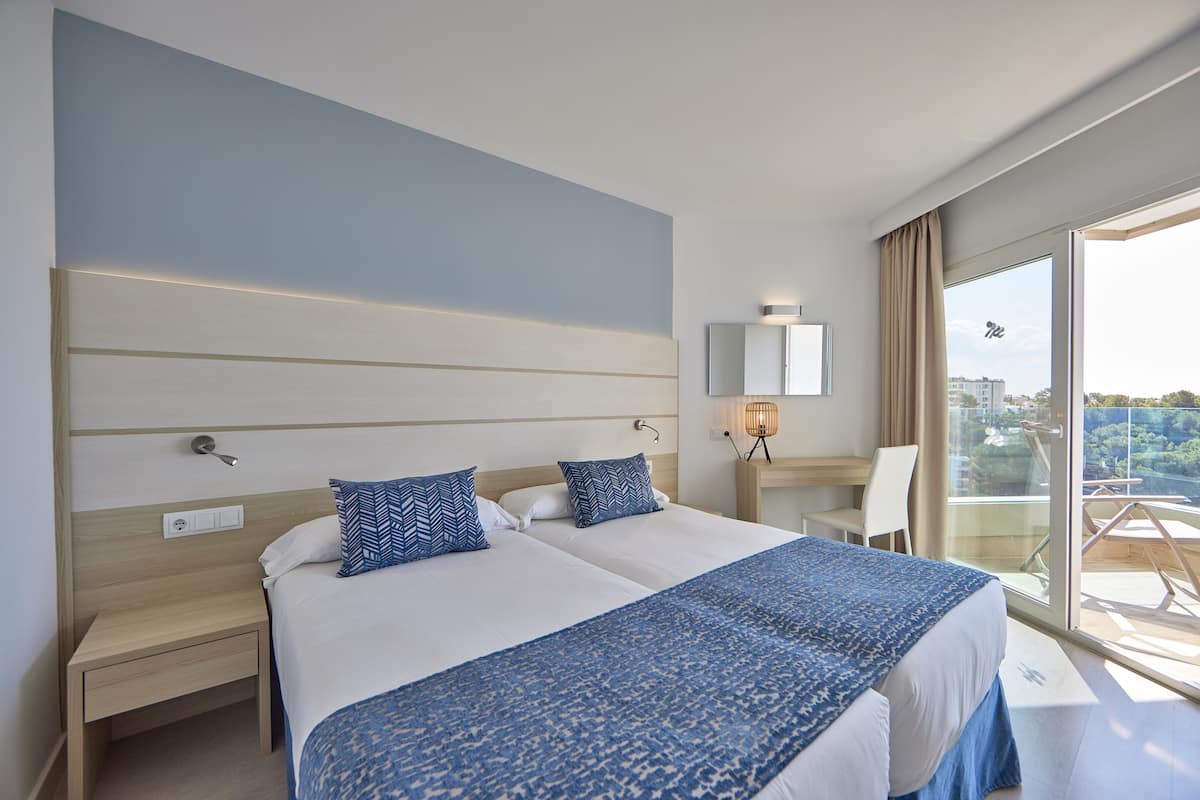 A hotel room with two beds, a desk, a chair, a large window with a view, and a balcony.