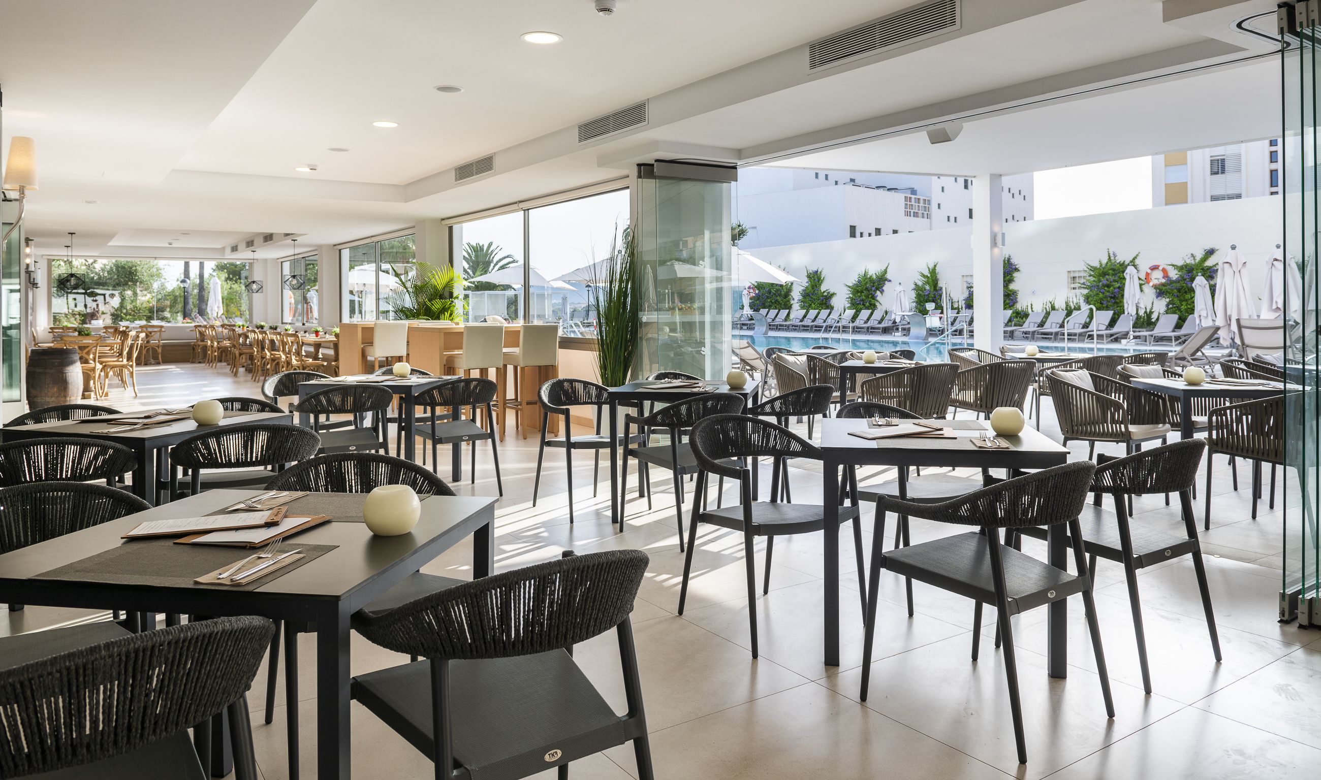 Un restaurante moderno con sillas y mesas de mimbre negro, ventanas amplias y vista a una piscina.