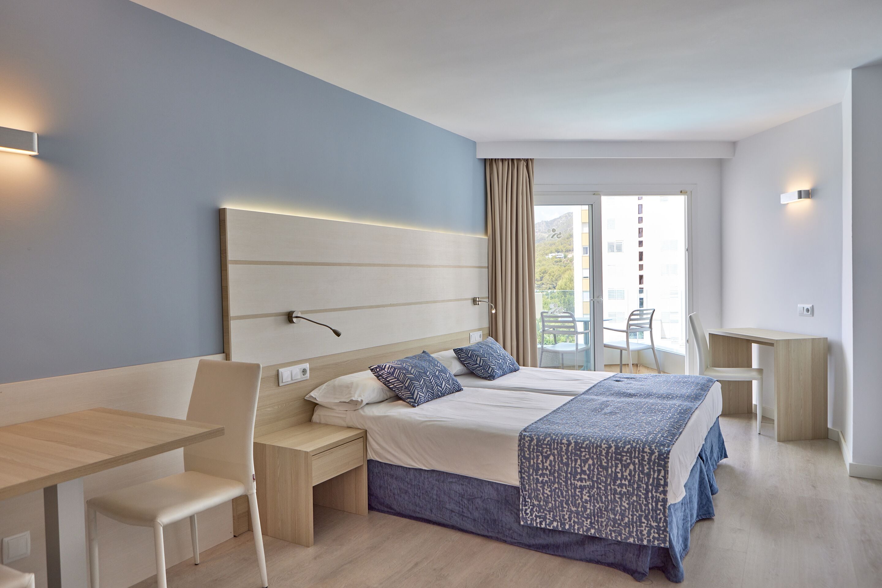 A modern hotel room with a large bed, a desk, and a chair. There is a window with curtains, and a balcony with chairs.