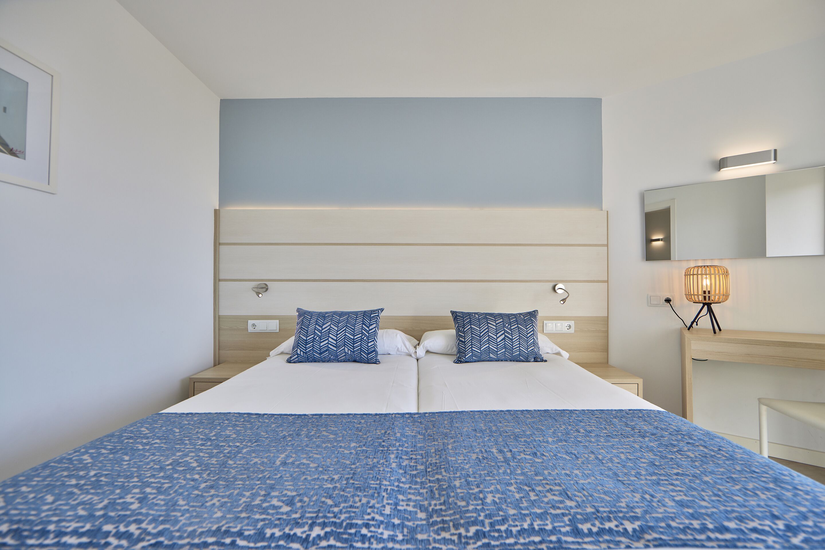 A double bed with a patterned blue bedspread, two matching pillows, a bedside table with a lamp, and a mirror on the wall.