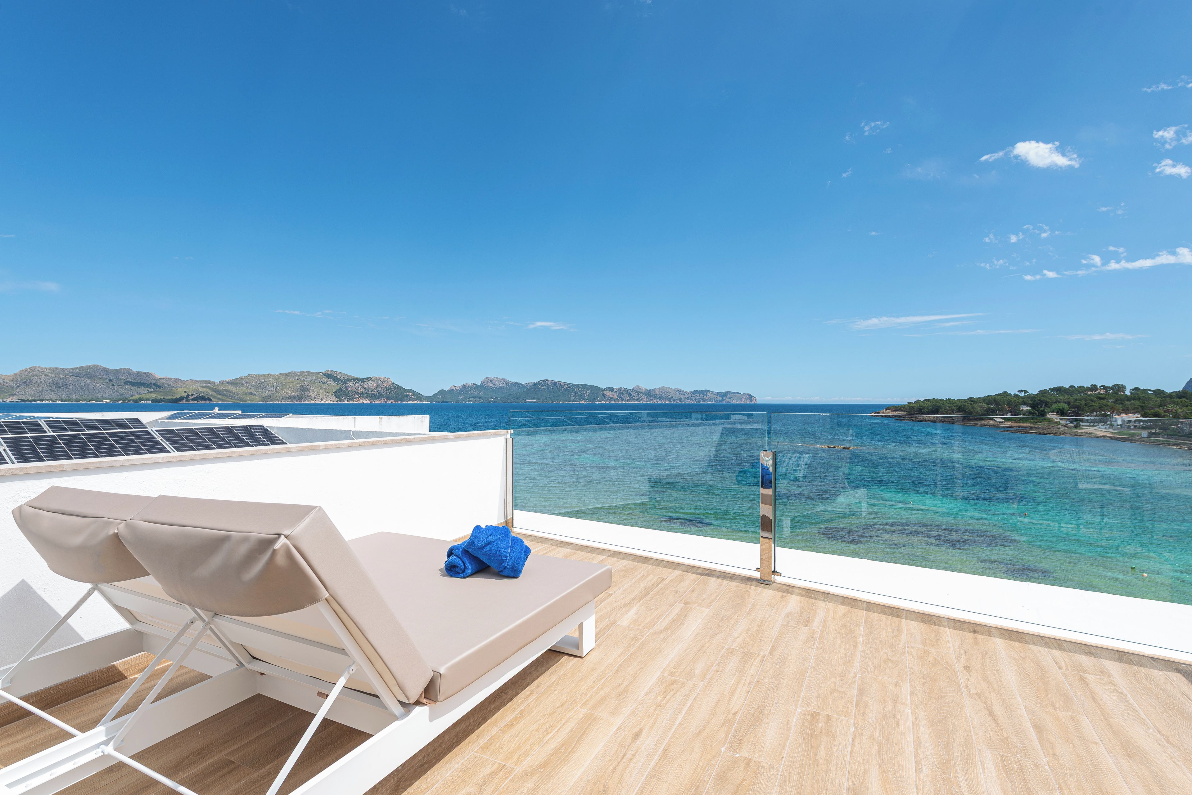 A sunlit balcony with a lounge chair and a towel, overlooking a clear blue sea and distant mountains.