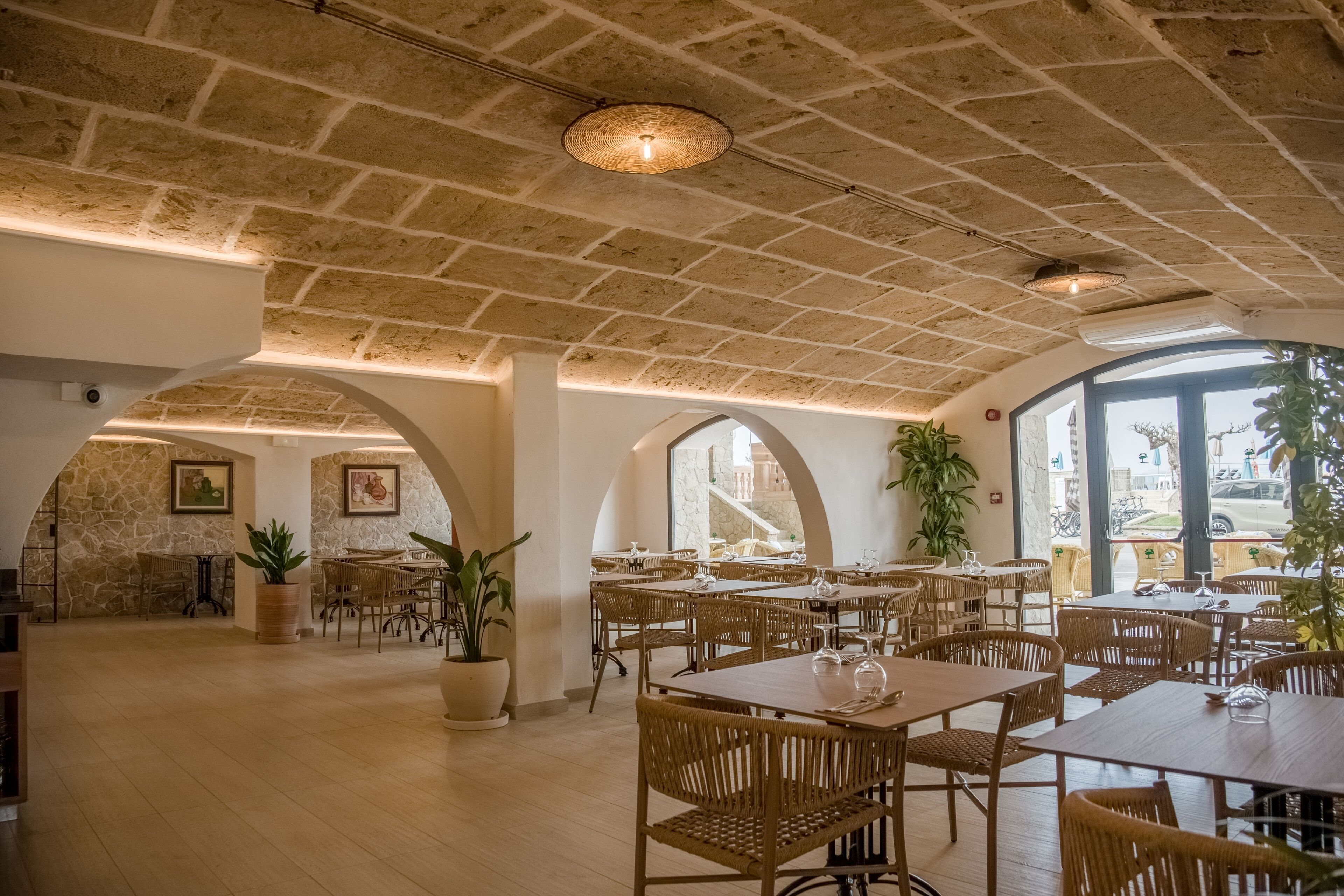 A spacious dining area with high ceilings, stone walls, and large windows. Tables are arranged in a U-shape, surrounded by wicker chairs.