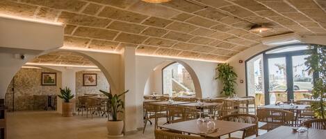 A spacious dining area with high ceilings, stone walls, and large windows. Tables are arranged in a U-shape, surrounded by wicker chairs.