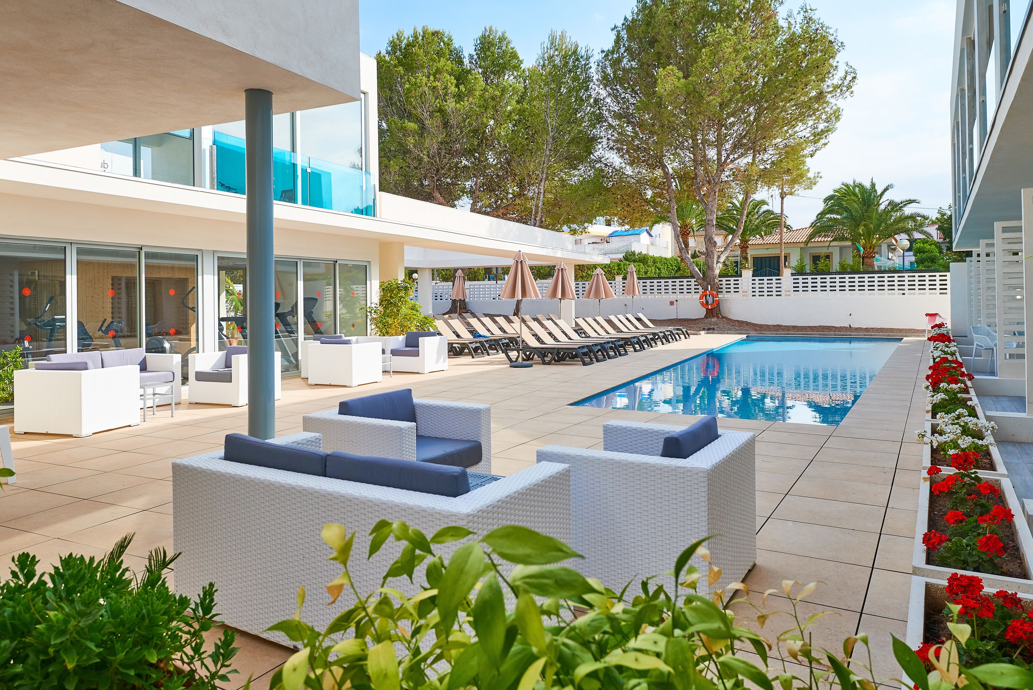 Un área de piscina moderna al aire libre con mobiliario de mimbre blanco, una piscina azul cristalina y flores rojas a lo largo del borde.