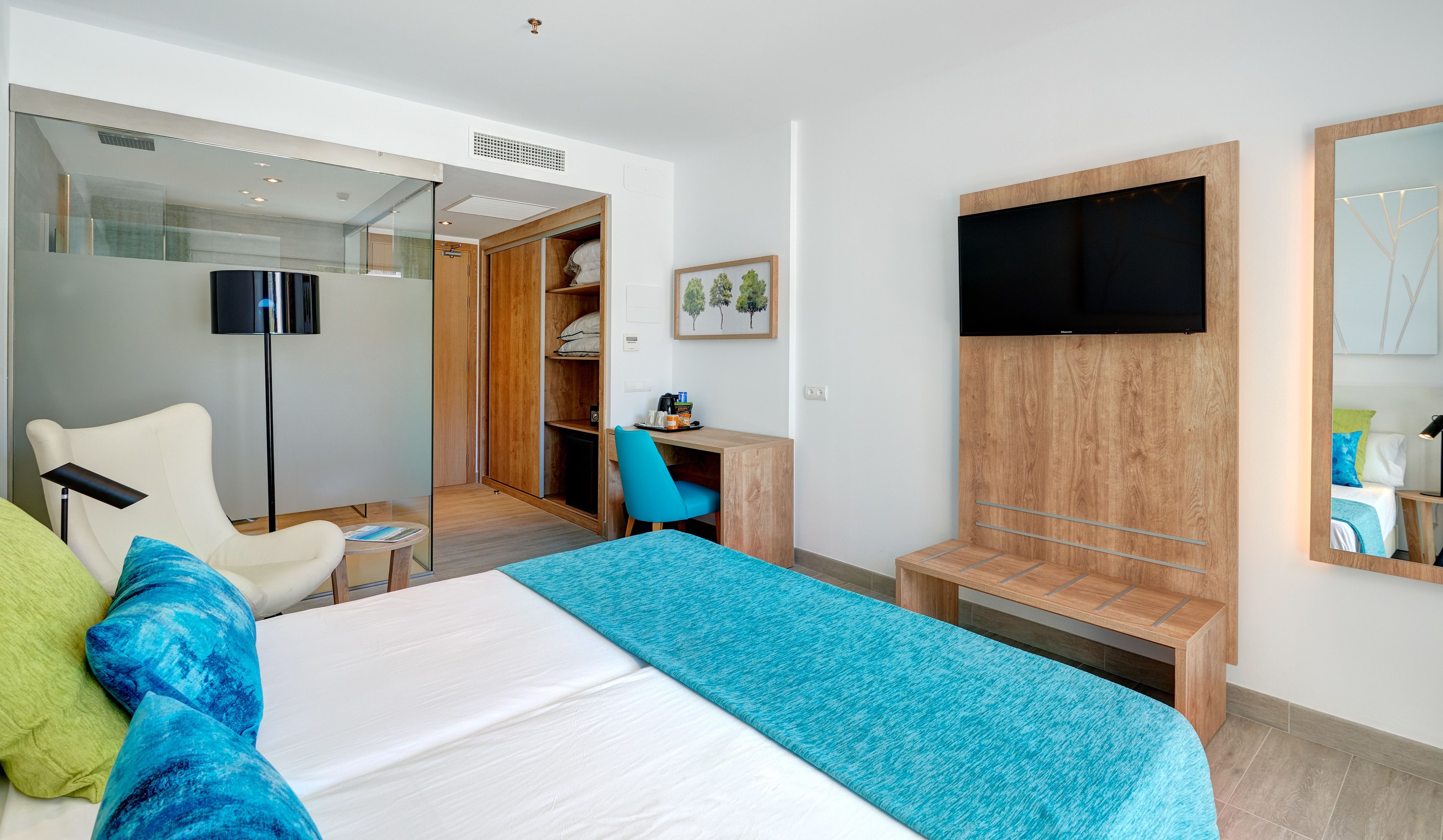 A modern hotel room with a bed, a desk, a chair, a wardrobe, and a television.