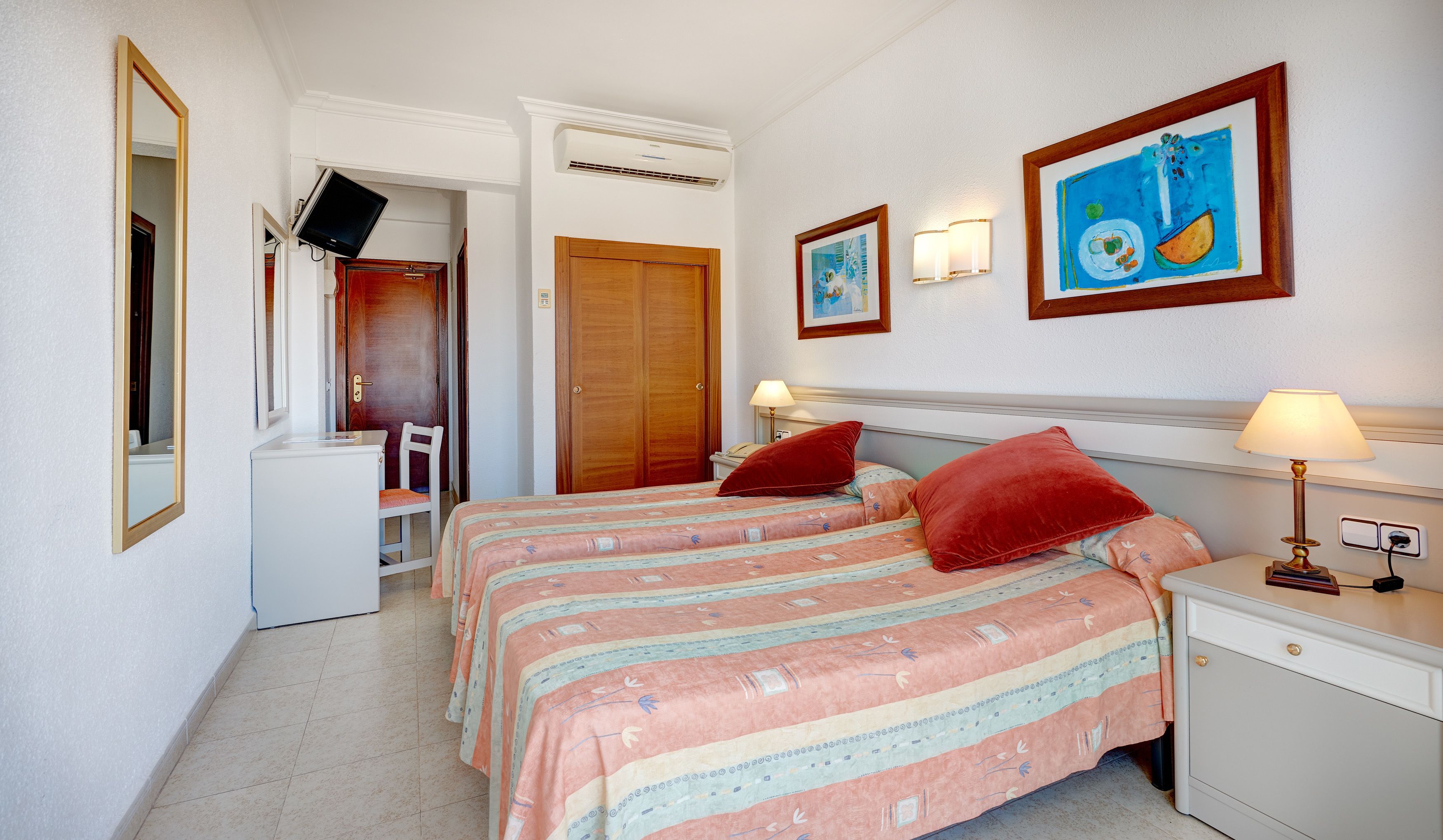 Una habitación de hotel con dos camas, un escritorio, una silla, un televisor y obras de arte en las paredes.