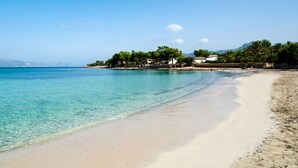 Una playa con aguas turquesas cristalinas, una orilla arenosa y un pequeño grupo de edificios con árboles.