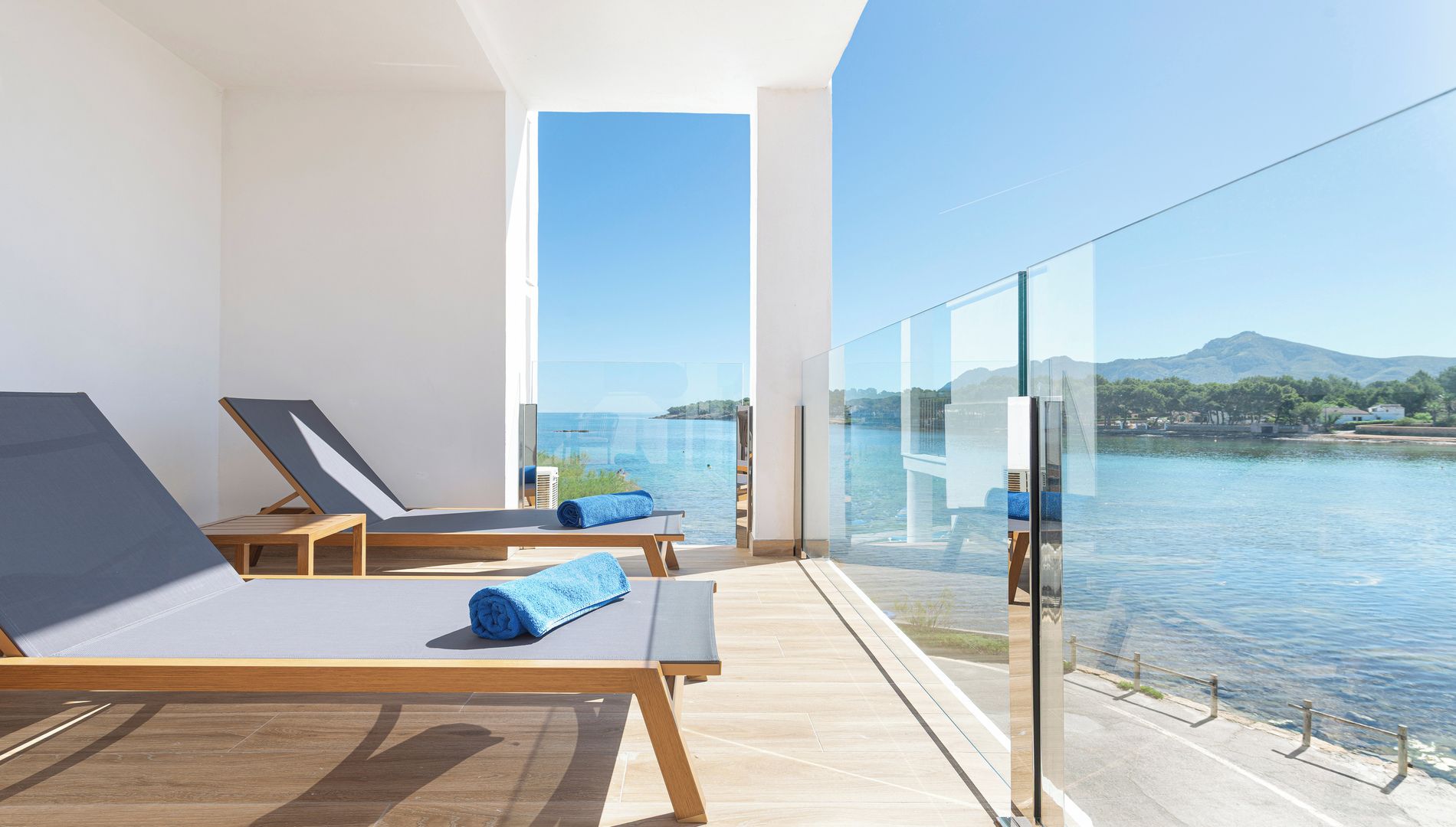 A modern balcony with sun loungers and a glass railing overlooking a body of water and distant landscape.