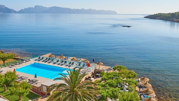 Zona de piscina con sillones de descanso y sombrillas, con vistas a una masa de agua tranquila y montañas al fondo.