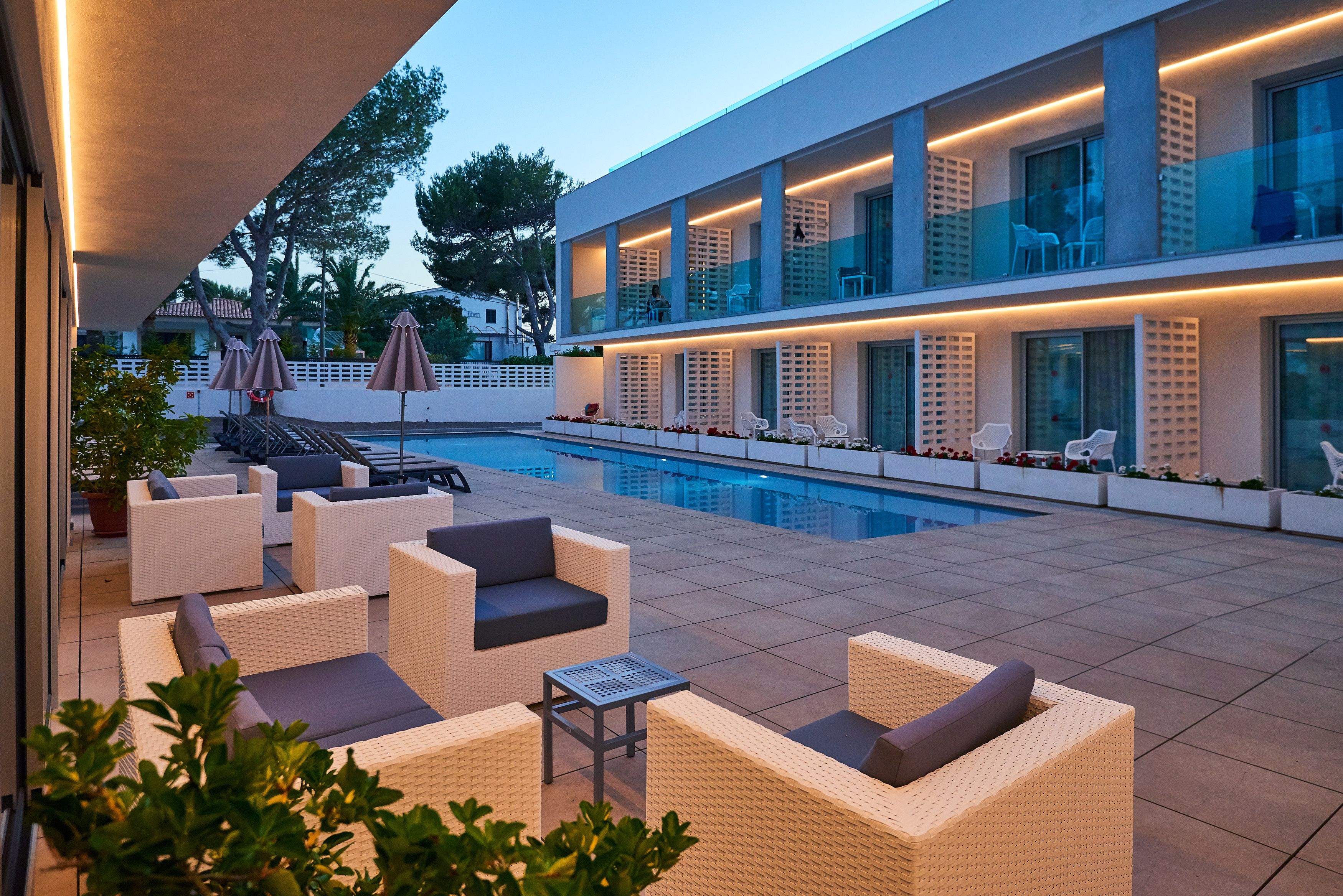 A modern outdoor pool area with lounge chairs and umbrellas, a swimming pool, and a building with large windows and balconies.