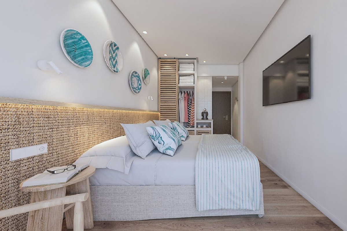 Un dormitorio moderno con una cabecera trenzada, una cama con ropa de cama a rayas, una mesita de noche con vasos y un libro, y un televisor montado en la pared.