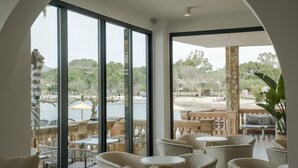 Amplio espacio interior con grandes ventanales que ofrecen vistas a una playa y zonas verdes. Hay varias áreas de estar con sillas y mesas.