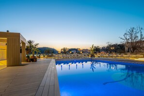 Piscina com espreguiçadeiras e plantas em vasos, cercada por um deck de madeira e um edifício com uma placa.