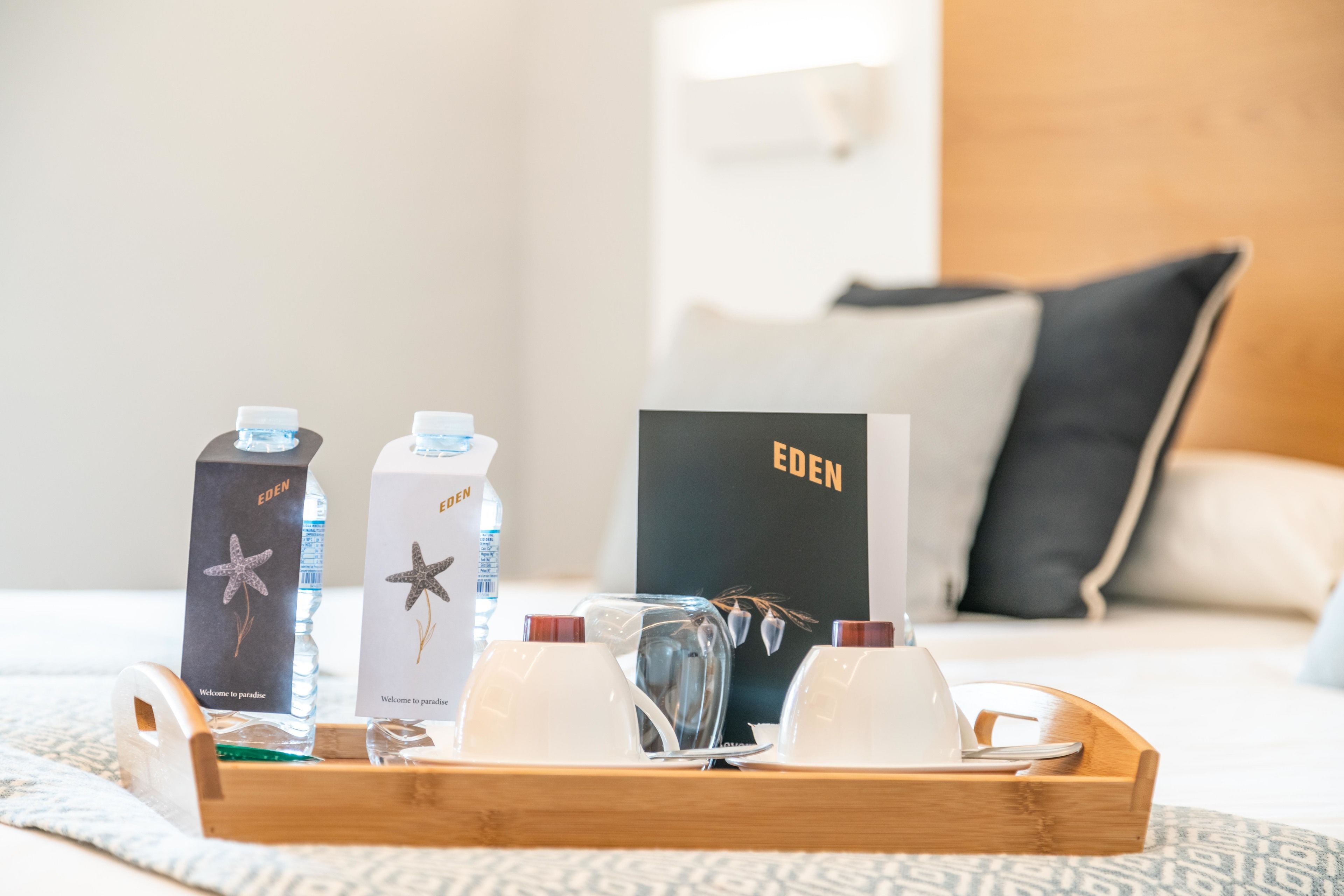 Een dienblad met een waterfles, een glas en een doosje met de tekst 'EDEN' op een bed met een decoratieve dekbedovertrek.